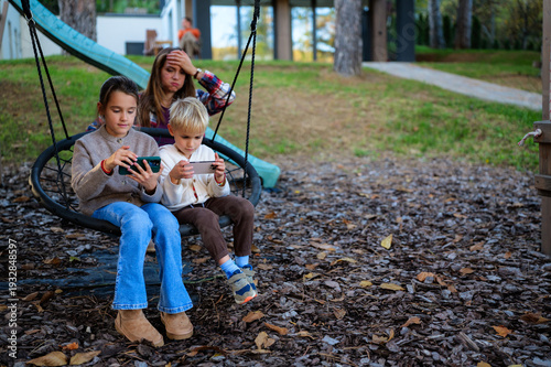 Bilde på lerret Two children on a swing absorbed in mobile games while their mother watches near