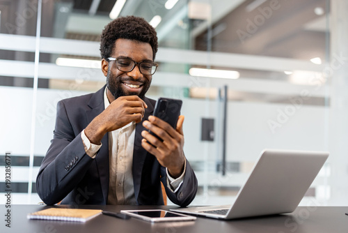Fotografie Smiling businessman browsing smartphone at a desk with open laptop in a modern o
