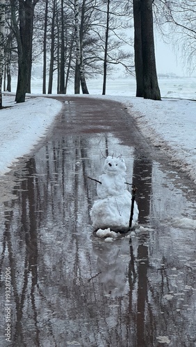 Konstfotografi Melting snowman with twig arms isolated in a large puddle on a slushy park path