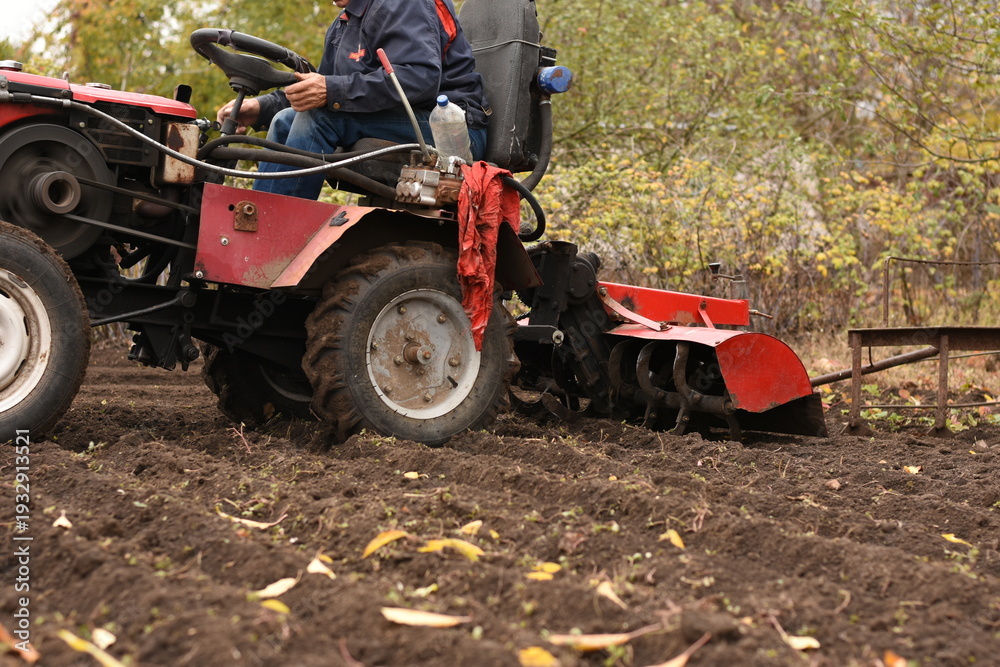 Obraz premium Man on red tractor plowing the field from side, medium shot, plow lowered into soil, horizontal shot.