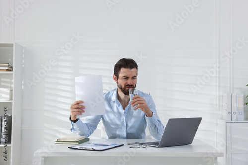 Man with glass of water suffering from heat at table near white wall indoors, space for text
