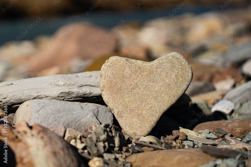 Obraz premium Natural beige river stone shaped like a heart lying on a rocky riverbank in bright sunlight with a blurred background of a mountain river. Symbol of love created by nature.