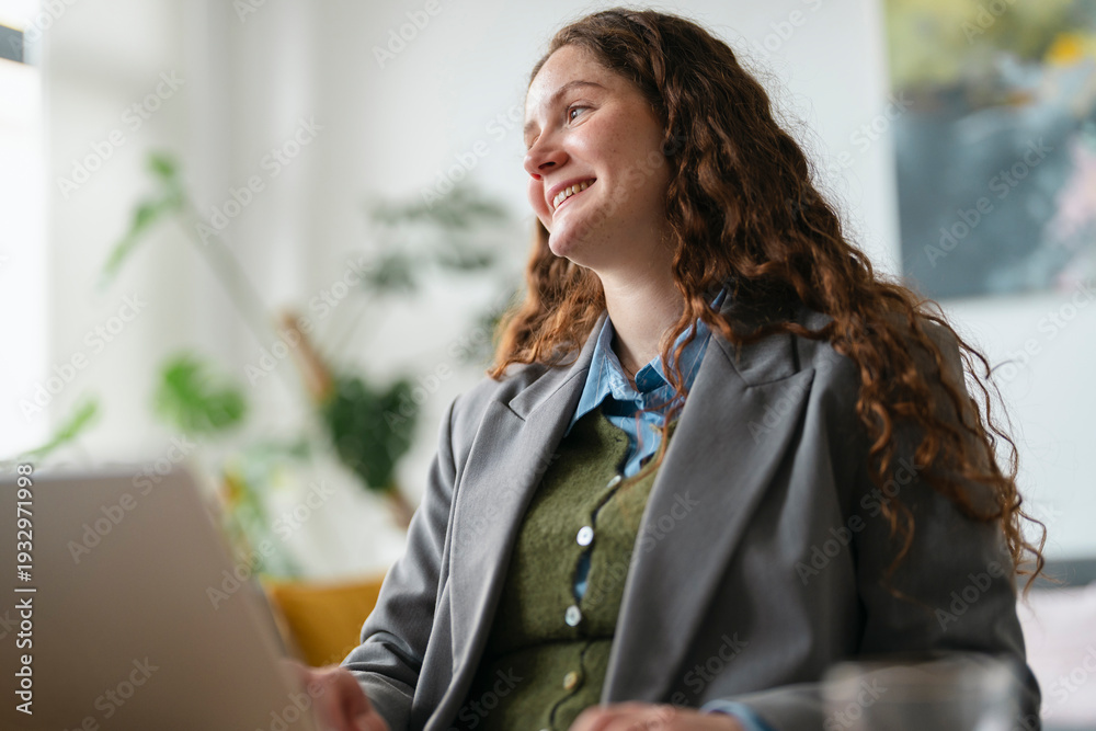 © Studio Marmellata - A young woman with curly brown hair smiles while working on her laptop in a bright, modern office setting.
