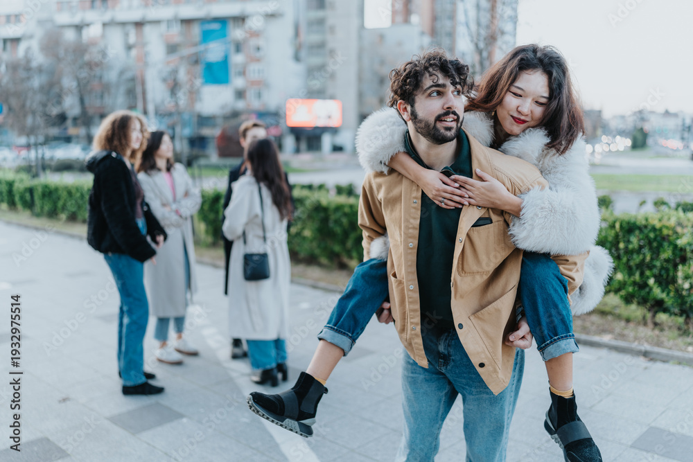 Fototapeta premium A man carries a woman on his back as they walk with friends in a city park, showing playful affection.
