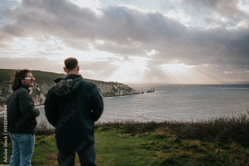 Fototapeta premium Two people standing on a grassy hill overlooking the ocean. The sky is cloudy and the sun is shining through the clouds