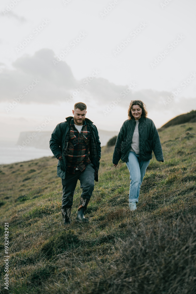 Fototapeta premium A man and woman are walking on a hillside. The man is wearing a plaid jacket and the woman is wearing a green jacket. The sky is cloudy and the couple is enjoying the view of the ocean