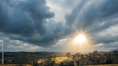 Sunbeams breaking through dramatic dark clouds over a vast rural landscape with rolling hills and trees casting long shadows