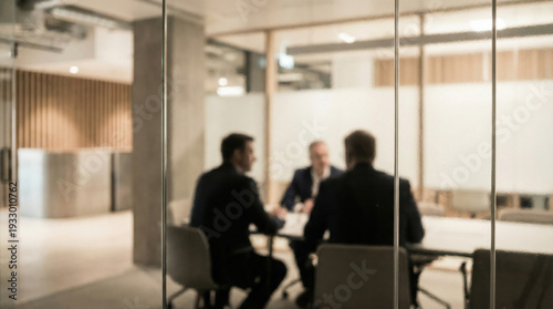 Three businessmen in suits are blurred and visible through glass meeting room doors during a discussion