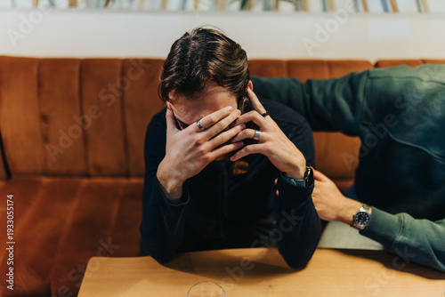 A man in a dark hoodie sits on a brown sofa with his hands over his face. A nearby friend reaches out, suggesting support in a casual living room scene.