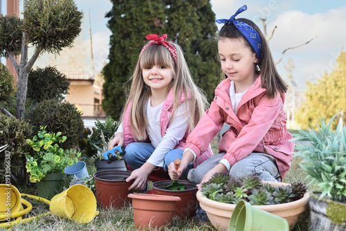 Two little girls planting flowers in garden pots outdoors on a sunny day. Children learning about nature, gardening and environmental care. Spring activity, childhood education and healthy lifestyle 