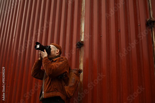 Fotografia A professional male photographer in a brown corduroy jacket and beanie captures photos with a DSLR camera against a tall, red corrugated metal industrial wall, holding a leather backpack