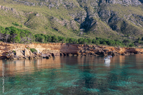 Calm turquoise sea and golden cliffs at Playa Es Calo in Mallorca with anchored boats and pine forest