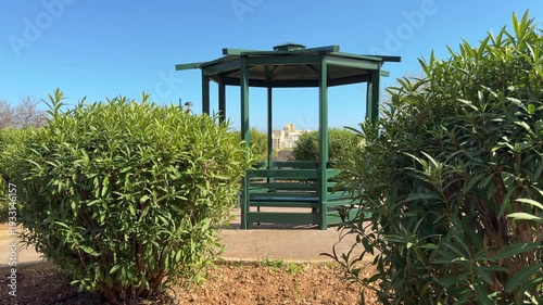 Classical dark green wooden gazebo with built-in benches in a peaceful public garden. Surrounded by Mediterranean plants, stone paving, and a clear blue sky. 