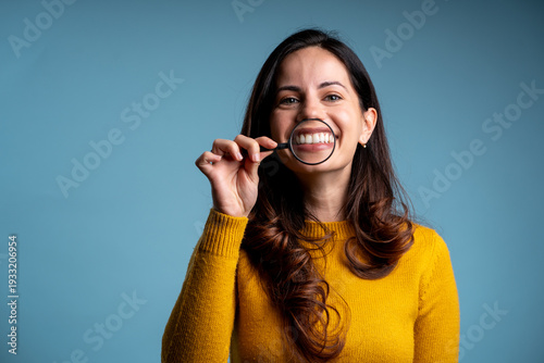 Happy female looking at camera, holding magnifying glass over perfect smile on blue