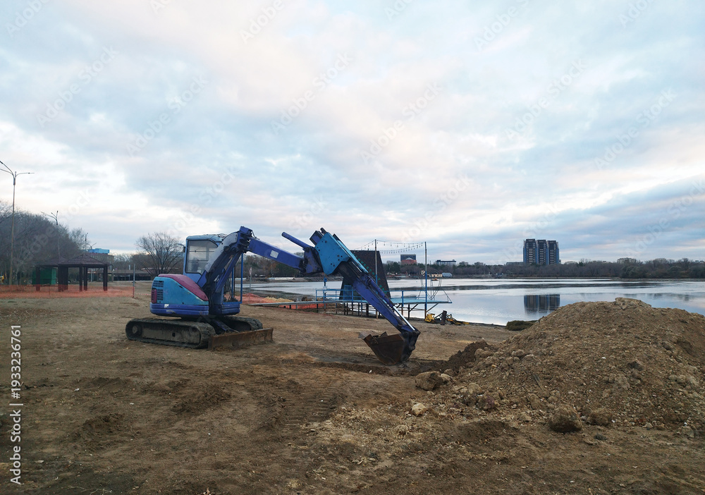 Fototapeta premium A small blue and purple excavator sits on muddy, brown ground next to a calm body of water against a wide, cloudy sky at dusk or on an overcast day.