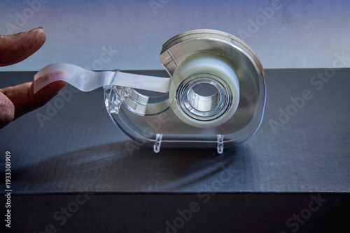 Close-up of a hand pulling transparent adhesive tape from a desktop tape dispenser on a dark surface