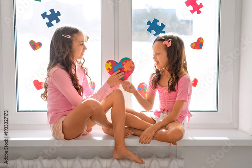 A girl gives a heart-shaped postcard to sister with an autism spectrum disorder. The window of the children's room is decorated with puzzle pieces.