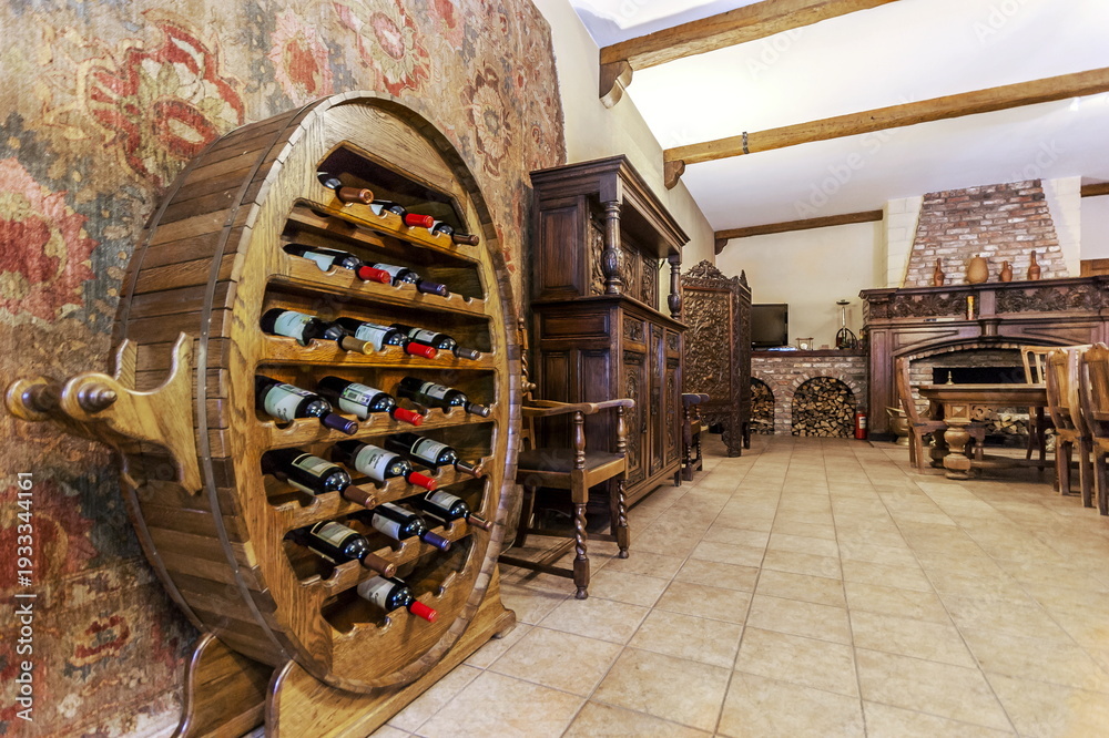 Fototapeta premium A wooden wine rack holds several bottles in a cozy room. The space features wooden furniture, a fireplace, and a decorative wall. The warm lighting creates a welcoming feel in the evening
