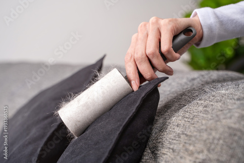 Close-up female hand using adhesive lint roller on black cushion, gray sofa removing pet hair. Concept of textile hygiene and maintaining clean living space through regular household cleaning routine