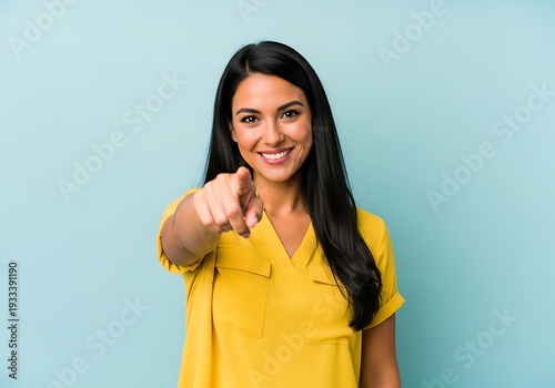 Smiling woman pointing directly at the camera with a playful expression on her face standing against a light blue background