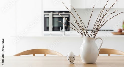 Willow catkins in a white pitcher beside an egg cup on a wooden table. Quiet luxury minimalist Easter table decoration concept.