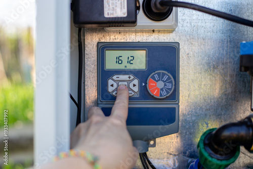A hand operates a digital irrigation timer on a control panel in a garden. smart farming, automated watering, modern technology.