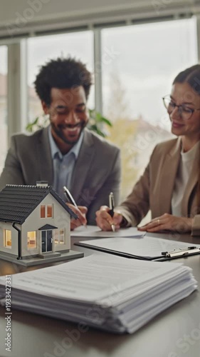 There's a model of a house on the table, and a realtor and a buyer are signing a sales contract nearby.
