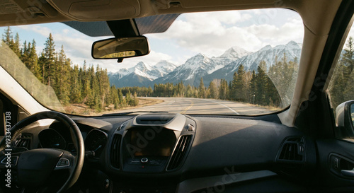 The view from inside a car driving on a scenic highway, looking through the windshield at lush pine forests and majestic snow-capped mountains under a bright sky.