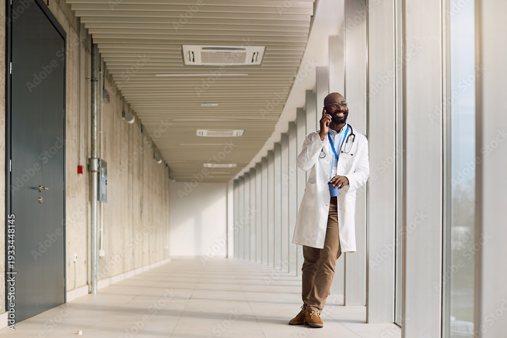 Fototapeta premium Happy black doctor talking on cell phone in hospital hallway.
