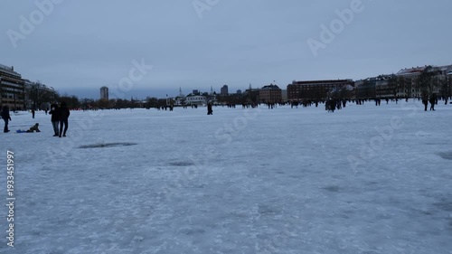 A winter landscape of Copenhagen, Denmark. The photograph depicts a wide, snow-covered square, or frozen urban space, in the center of the capital, where people stroll on a cold winter day