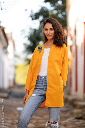 Woman poses on a cobblestone street in a colorful town wearing a bright yellow coat and casual jeans during a sunny day