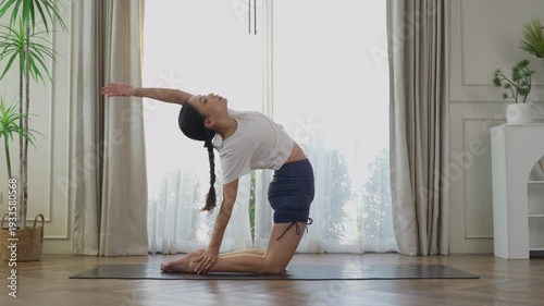 Young Asian woman practicing camel yoga pose on mat in bright living room, healthy lifestyle home workout stretching flexibility mindfulness wellness