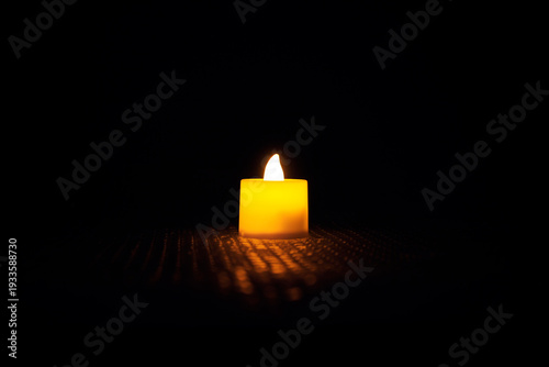 Small LED candle with soft warm flame in dark black background creating dramatic low light atmosphere. Minimalist night photography symbolizing hope, peace, faith, prayer, dramatic black backdrop