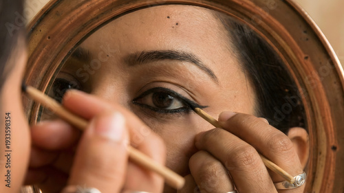 A close-up reflection in a polished bronze mirror of a woman applying kohl to her eyes with a small stick