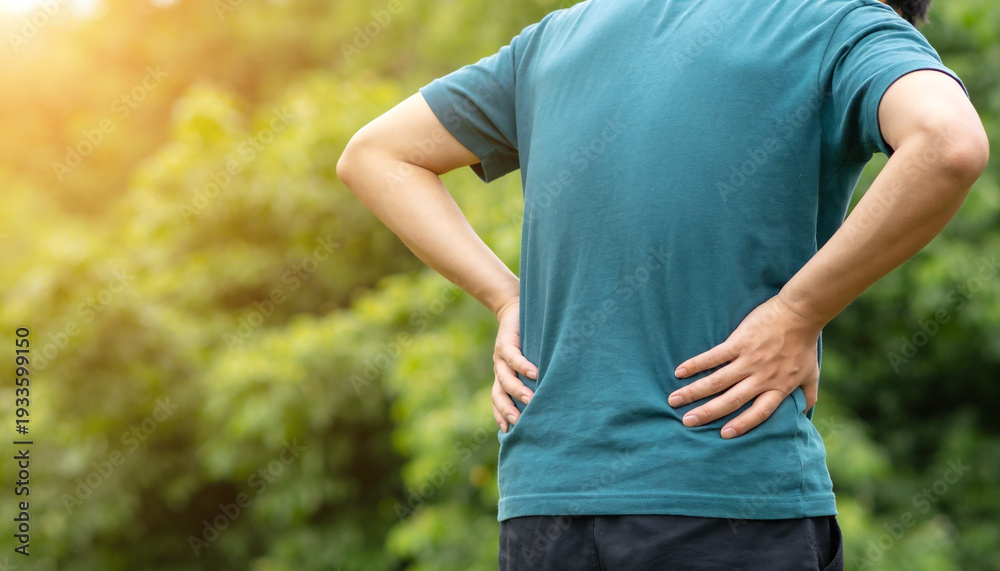 Naklejka premium Person holding aching lower back with hands outdoors with blurred green foliage background