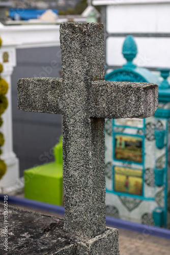 Stone cross stands at a cemetery with colorful tombs and a clear sky in the background during daylight hours