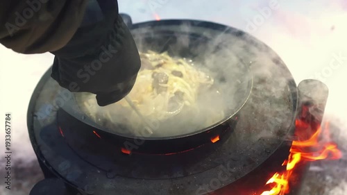 Hand stirring hot meal over wood burning stove in cold weather. Camping adventure concept, outdoor cuisine and cozy winter mood. Pilaf with beef meat and rice boiling.