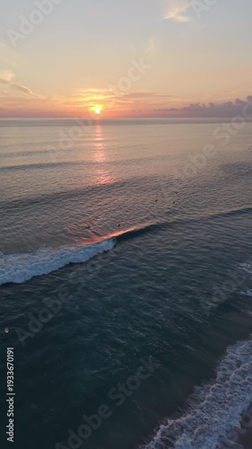 Vertical aerial pullback at tropical sunset revealing surfers and palm trees along ocean coastline