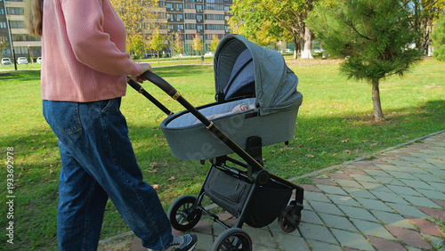 A young woman enjoys a relaxing stroll with her newborn baby in a stroller. Scene set in a city park with green grass and autumn trees on a bright day. Lifestyle concept of parenting and leisure