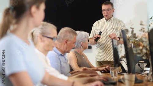 Enthusiastic male tutor teaching group of seniors in computer workshop, promoting technology adoption among older generations