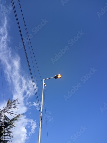 Street lamp illuminated against a clear blue sky with wispy clouds and overhead wires, capturing a moment of urban illumination in the daytime