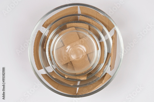 A top down view of three clear glass bowls of different sizes nested together with brown cardboard pieces used as protective spacers on a white background