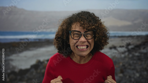 Foto Woman in bright red sweater clenches fists on rocky beach as waves crash behind; determination resilience