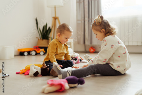 Two children sit on the floor surrounded by toys. They spend time playing and interacting with each other. Sunlight fills the bright room, creating a lively scene