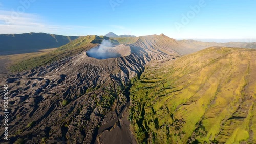 Cinematic aerial view of active Mount Bromo volcano emitting smoke with Mount Semeru in the distance at sunrise. Dramatic volcanic landscape in East Java, Indonesia.