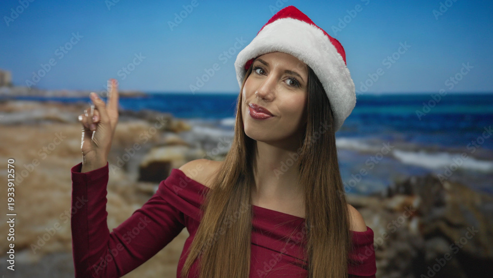 Fototapeta premium Woman wearing santa hat and burgundy top pointing finger to sky at sunny beach shore under clear blue sky; anticipation.