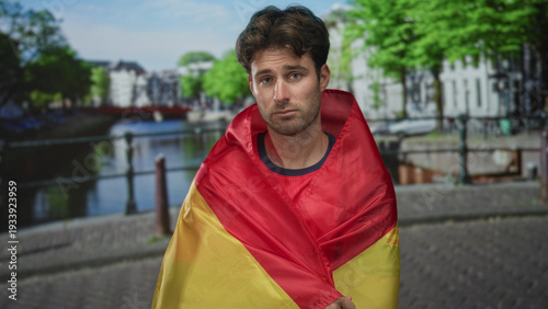 Young hispanic man wrapped in a red and yellow flag on a cobblestone street by amsterdam canal; melancholy reflection.