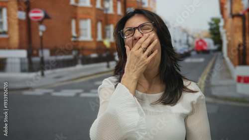 Картината върху платно Hispanic woman wearing white blouse and glasses covering mouth with hand on city street; fatigue exhaustion