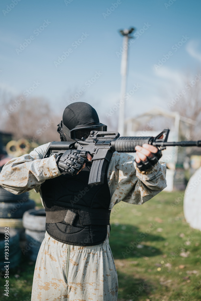 Fototapeta premium Paintball player in camouflage gear aims a rifle during an outdoor game. The scene shows a tactical athlete on a field with barriers and tires in the background.