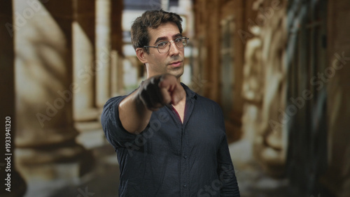 Foto Man points finger forward while wearing glasses and a dark shirt in a columned building corridor; confidence challenge